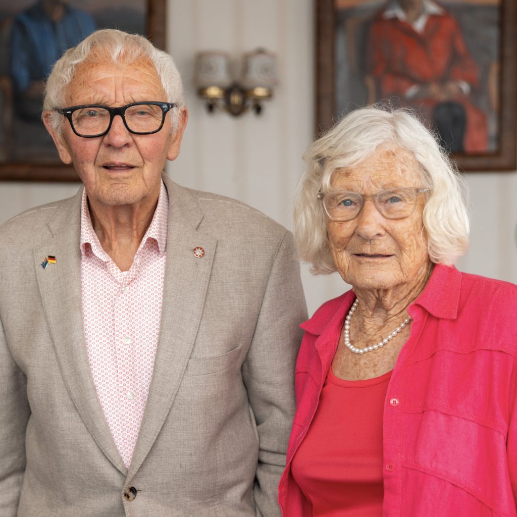 2025 Kiwibank New Zealander of the Year Semi-Finalists Sir Mark Dunajtschik and Dorothy Spotswood, photographed indoors. Sir Mark has shirt white hair, and wears blue glasses, a tan blazer, and pink shirt. Dorothy has length white hair, and wears a pink shirt open over a red top, and pearls.